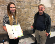 Eine Frau mit einem aufgeschlagenen Ordner steht neben einem lächelnden Mann vor einer Steinwand mit Vitrinen, die verschiedene Objekte enthalten, in der Hochschule Coburg. Beide wirken zufrieden und lässig in ihrer Kleidung.