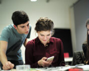 Drei junge Leute sitzen um einen Tisch in einem Studienraum der Hochschule Coburg. Einer steht und beugt sich vor, zwei sitzen. Die Person in der Mitte schaut auf ein Smartphone, die anderen schauen aufmerksam zu.