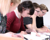 Vier Studierende sitzen in einer Reihe an einem Tisch und sind in ihre Studien vertieft. Sie machen sich Notizen und lesen aus den vor ihnen ausgebreiteten Materialien vor. Sie verkörpern damit die Hingabe, die man an der Hochschule Coburg oft sieht. Die Umgebung erinnert an ein Klassenzimmer oder eine Lerngruppe.