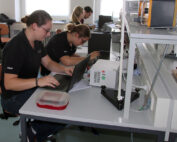 In einem modernen Labor der Hochschule Coburg arbeiten die Menschen fleißig an Schreibtischen und konzentrieren sich auf Laptops und wissenschaftliche Geräte. Der Raum ist hell erleuchtet, mit eleganten Möbeln und verschiedenen Instrumenten, über die Tische spannen sich Kabel.