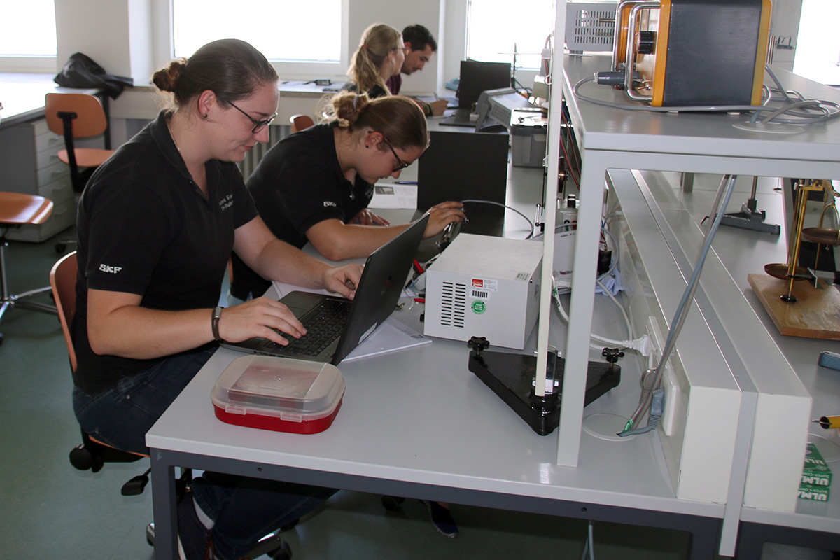 In einem modernen Labor der Hochschule Coburg arbeiten die Menschen fleißig an Schreibtischen und konzentrieren sich auf Laptops und wissenschaftliche Geräte. Der Raum ist hell erleuchtet, mit eleganten Möbeln und verschiedenen Instrumenten, über die Tische spannen sich Kabel.