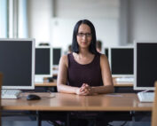 Eine Frau mit Brille und langen dunklen Haaren sitzt an einem Schreibtisch und blickt in die Kamera. Sie ist umgeben von Computermonitoren in einem hellen, modernen Büro im hochmodernen Computerlabor der Hochschule Coburg.