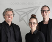 Drei lächelnde Menschen in schwarzer Kleidung stehen vor einer Wand mit dem Logo „COBURGER DESIGNPILOT“. Das Design eines Papierfliegers steht für den Innovationsgeist der Hochschule Coburg.