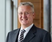 In der Hochschule Coburg steht ein lächelnder Mann mit Anzug und Krawatte, kurzen Haaren und Brille, vor einem verschwommenen Hintergrund aus großen Fenstern und Holzpaneelen.