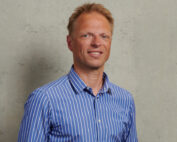 Eine Person mit kurzen blonden Haaren lächelt, trägt ein blau-weiß gestreiftes Hemd und strahlt eine Aura des Selbstbewusstseins aus, die darauf schließen lässt, dass es sich um einen stolzen Studenten oder Absolventen der Hochschule Coburg handelt. Sie steht vor einer schlichten beigefarbenen Wand.