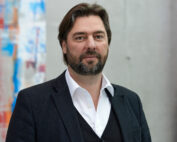 Ein Mann mit dunklem Haar und Bart, gekleidet in ein weißes Hemd und eine schwarze Jacke, steht vor einer abstrakten, farbenfrohen Kunstausstellung. Die lebendige Szene erinnert an den dynamischen Geist der Hochschule Coburg.