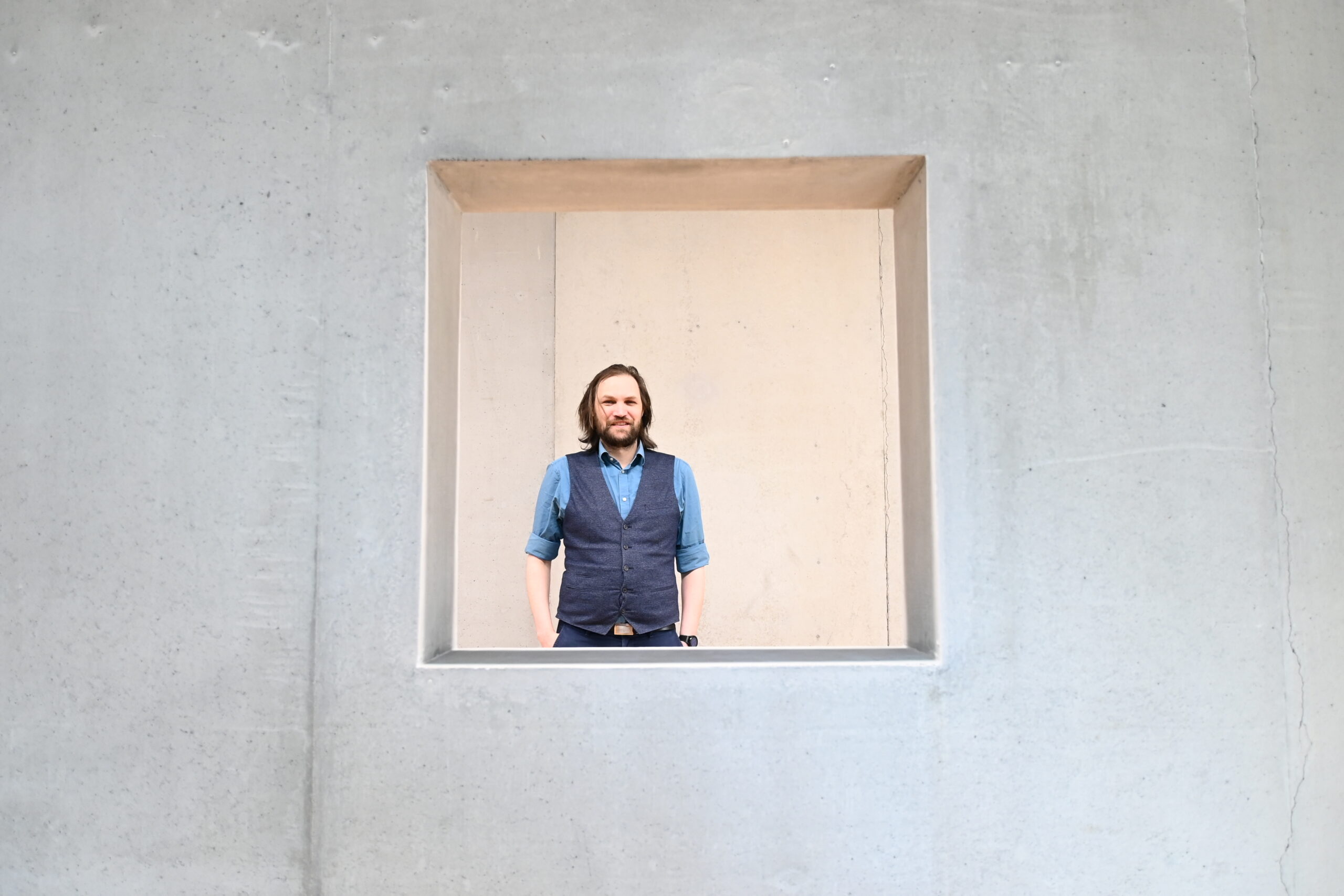 In einem quadratischen Fensterrahmen der Hochschule Coburg steht ein Mann mit langen Haaren und Bart. Er trägt ein blaues Hemd und eine dunkle Weste, vor einer Betonwand.