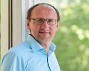 Eine Person mit Brille und hellblauem Hemd steht an einem großen Fenster der Hochschule Coburg. Sie hat kurze Haare und einen neutralen Gesichtsausdruck. Im Hintergrund ist verschwommenes Grün draußen zu sehen, was eine angenehme Außenumgebung suggeriert.