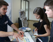 Drei junge Erwachsene sitzen an einem Tisch der Hochschule Coburg und diskutieren über Drucksachen. Einer hält einen Vortrag, die anderen beiden hören aufmerksam zu. Auf dem Tisch und in einem Ständer in der Nähe liegen Broschüren und Prospekte der Hochschule.
