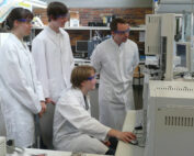 In einem geschäftigen Labor der Hochschule Coburg arbeiten vier Wissenschaftler in weißen Laborkitteln und Schutzbrillen konzentriert zusammen. Einer sitzt an einem Computer, während die anderen stehen und das komplexe Zusammenspiel der wissenschaftlichen Instrumente und Geräte um sie herum beobachten.