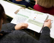 In einem Hörsaal der Hochschule Coburg sitzt eine Person mit einem aufgeschlagenen Buch in Deutsch auf dem Schoß, in dem ein Wortsuchrätsel steht. Die Mitschüler in der Nähe konzentrieren sich auf ihre Studien, während auf dem Tisch neben ihnen ein Notizblock liegt.