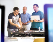 Drei Männer sitzen um einen Tisch an der Hochschule Coburg und arbeiten an einem technischen Projekt. Einer stellt ein Gerät ein, ein anderer hält einen Laptop und der dritte beobachtet. Kabel und Geräte liegen verstreut auf dem Tisch, im Vordergrund sind unscharfe Bildschirme zu sehen, die innovative Vorhaben einfangen.