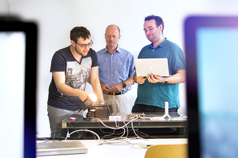 Drei Männer sitzen um einen Tisch an der Hochschule Coburg und arbeiten an einem technischen Projekt. Einer stellt ein Gerät ein, ein anderer hält einen Laptop und der dritte beobachtet. Kabel und Geräte liegen verstreut auf dem Tisch, im Vordergrund sind unscharfe Bildschirme zu sehen, die innovative Vorhaben einfangen.