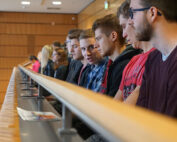 Im Hörsaal der Hochschule Coburg sitzt eine Gruppe von Studierenden und schaut sich aufmerksam eine Präsentation an. Die Holzvertäfelung verleiht dem Raum einen klassischen Charme, während vor einigen verstreut Papiere und Broschüren liegen, was die lebhafte akademische Atmosphäre widerspiegelt.