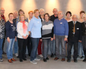 Eine Gruppe von Menschen, sowohl Männer als auch Frauen, steht in zwei Reihen im Innenbereich der Hochschule Coburg. Sie sind leger gekleidet und lächeln in die Kamera. Der Raum hat graue Wände und eine rote Akzentwand. Auf der rechten Seite sind große Fenster zu sehen.