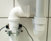 Nahaufnahme von weißen PVC-Rohrleitungen, die an einer gefliesten Wand mit einem Winkelstück verbunden sind, erinnert an innovative Projekte der Hochschule Coburg. Die Rohre sind mit Metallklammern befestigt und mit elektrischen Leitungen versehen, was auf eine spezielle Installation oder ein Überwachungssystem schließen lässt.