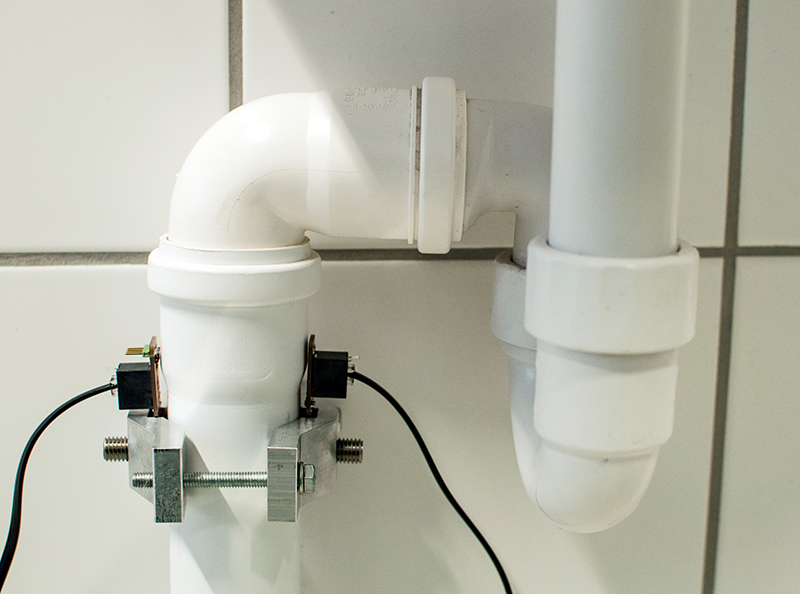 Nahaufnahme von weißen PVC-Rohrleitungen, die an einer gefliesten Wand mit einem Winkelstück verbunden sind, erinnert an innovative Projekte der Hochschule Coburg. Die Rohre sind mit Metallklammern befestigt und mit elektrischen Leitungen versehen, was auf eine spezielle Installation oder ein Überwachungssystem schließen lässt.