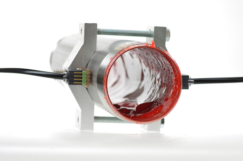 Ein metallisches, zylindrisches Gerät, das in einen Ständer eingespannt ist und einem Projekt aus dem Advanced Engineering-Programm der Hochschule Coburg ähnelt, weist an den Seiten angebrachte Drähte auf. Ein Ende des Zylinders ist offen und gibt den Blick auf eine rote, gelartige Substanz im Inneren frei.