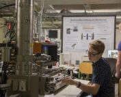 Eine Person mit Brille bedient ein großes wissenschaftliches Instrument in einem Labor der Hochschule Coburg. Sie sitzt da und arbeitet konzentriert an den Bedienelementen. Hinter ihr hängt ein Poster mit Texten und Diagrammen, während die Umgebung mit verschiedenen Geräten und Kabeln vollgestopft ist.