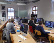 In einem Hörsaal der Hochschule Coburg sitzen die Studierenden an Tischen mit Apple-Computern. Ein Lehrer steht neben Whiteboard und Projektionsleinwand und leitet die Sitzung. Der Raum wird durch große Fenster erhellt und mit Schwarzweißfotos geschmückt.