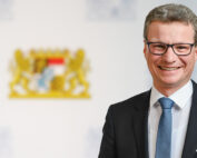 Ein Mann in Anzug und Krawatte lächelt in die Kamera, seine Brille zeugt von Intellekt. Selbstbewusst steht er vor einem unscharfen Emblem auf weißem Grund, das die Hochschule Coburg repräsentiert.