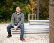 Eine Person mit Brille, grauem Hemd, blauer Jeans und roten Schuhen sitzt auf einer modernen, mehrstufigen Holzbank in der Parkanlage nahe der Hochschule Coburg mit Bäumen im Hintergrund.