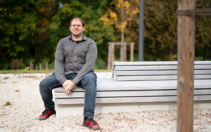 Eine Person mit Brille, grauem Hemd, blauer Jeans und roten Schuhen sitzt auf einer modernen, mehrstufigen Holzbank in der Parkanlage nahe der Hochschule Coburg mit Bäumen im Hintergrund.
