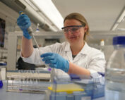 Eine Wissenschaftlerin der Hochschule Coburg, mit Schutzbrille und Laborkittel, benutzt im Labor eine Pipette. Sie trägt blaue Handschuhe und ist von einer Reihe von Laborgeräten und Flaschen umgeben.