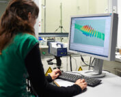 Eine Person im grünen Hemd sitzt an einem Schreibtisch und bedient einen Computer im Labor der Hochschule Coburg. Der Monitor zeigt farbenfrohe 3D-Grafiken oder Datenvisualisierungen, während daneben ein wissenschaftliches Instrument steht. Im Hintergrund sind verschiedene Laborgeräte zu sehen.