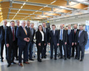 Eine Gruppe von fünfzehn Personen in Business-Kleidung steht in einer modernen Industrieanlage der Hochschule Coburg. Sie lächeln und posieren für das Foto vor Maschinen und Informationstafeln. Die Decke besteht aus Holzbögen und Industriebeleuchtung, was das anspruchsvolle Ambiente noch verstärkt.