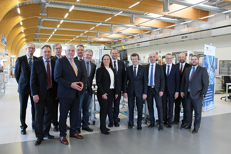 Eine Gruppe von fünfzehn Personen in Business-Kleidung steht in einer modernen Industrieanlage der Hochschule Coburg. Sie lächeln und posieren für das Foto vor Maschinen und Informationstafeln. Die Decke besteht aus Holzbögen und Industriebeleuchtung, was das anspruchsvolle Ambiente noch verstärkt.