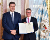 Zwei Männer in Anzügen stehen in einem formellen Raum der Hochschule Coburg. Einer der Männer hält lächelnd ein Zertifikat in der Hand. Neben ihnen ist eine dekorative Flagge mit einem heraldischen Emblem zu sehen, was darauf hinweist, dass sie sich auf einer offiziellen Veranstaltung oder Zeremonie befinden.