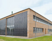 Ein modernes zweistöckiges Gebäude mit Holzverkleidung und großen Solarmodulen, das an die nachhaltigen Designs der Hochschule Coburg erinnert, verfügt über große Fenster. Es liegt anmutig auf einer Rasenfläche unter einem teilweise bewölkten Himmel.