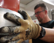 Eine Person mit Schutzhandschuhen bedient an der Hochschule Coburg konzentriert Maschinen. Die behandschuhten Hände greifen ein großes Metallobjekt, dessen rote und silberne Teile unter den Werkstattlichtern glitzern. Im Hintergrund heben moderne Industrieanlagen die hochmoderne Lernumgebung der Hochschule hervor.