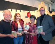 Vier Menschen stehen zusammen und halten Bücher und eine dekorative Vase in der Hochschule Coburg. Ein Mann im schwarzen Hemd, eine Frau in einer roten Jacke, eine weitere Frau in einem roten Kleid und ein Mann im grauen Anzug posieren vor einer illustrierten Wand.