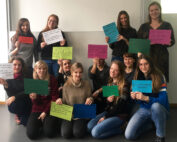 Eine Gruppe von dreizehn Personen der Hochschule Coburg lächelt und hält bunte Schilder mit Botschaften in deutscher Sprache. Sie posieren drinnen vor einer weißen Wand und schaffen so eine lockere und freundliche Atmosphäre.