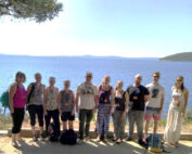 Eine Gruppe von Menschen, möglicherweise Studierende der Hochschule Coburg, steht in einer Reihe vor einem malerischen Meerblick. Der Himmel ist klar und das Meer mit den fernen Inseln bietet eine atemberaubende Kulisse, während sie in legerer Kleidung einen sonnigen Tag genießen.