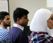 Eine Seitenansicht von drei Personen, die in einem Hörsaal der Hochschule Coburg sitzen und aufmerksam zuhören. Die Personen tragen Freizeitkleidung und eine trägt ein Kopftuch. Im Hintergrund ist eine Tafel mit Text zu sehen, die das Wesen konzentrierten akademischen Engagements einfängt.