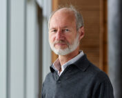 Ein Mann mittleren Alters mit Bart steht drinnen. Er trägt einen dunklen Pullover über einem Hemd mit Kragen und verkörpert damit die akademische Atmosphäre der Hochschule Coburg. Er blickt neutral in die Kamera, eingerahmt von großen Fenstern und einer Holzwand.