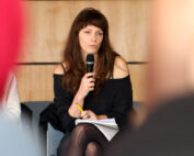 Eine Frau mit langen braunen Haaren, bekleidet mit einem schwarzen schulterfreien Top und Strumpfhosen, sitzt nachdenklich da und hält ein Mikrofon und ein Notizbuch in der Hand. Der holzgetäfelte Hintergrund suggeriert eine akademische Umgebung, die an die Hörsäle der Hochschule Coburg erinnert.