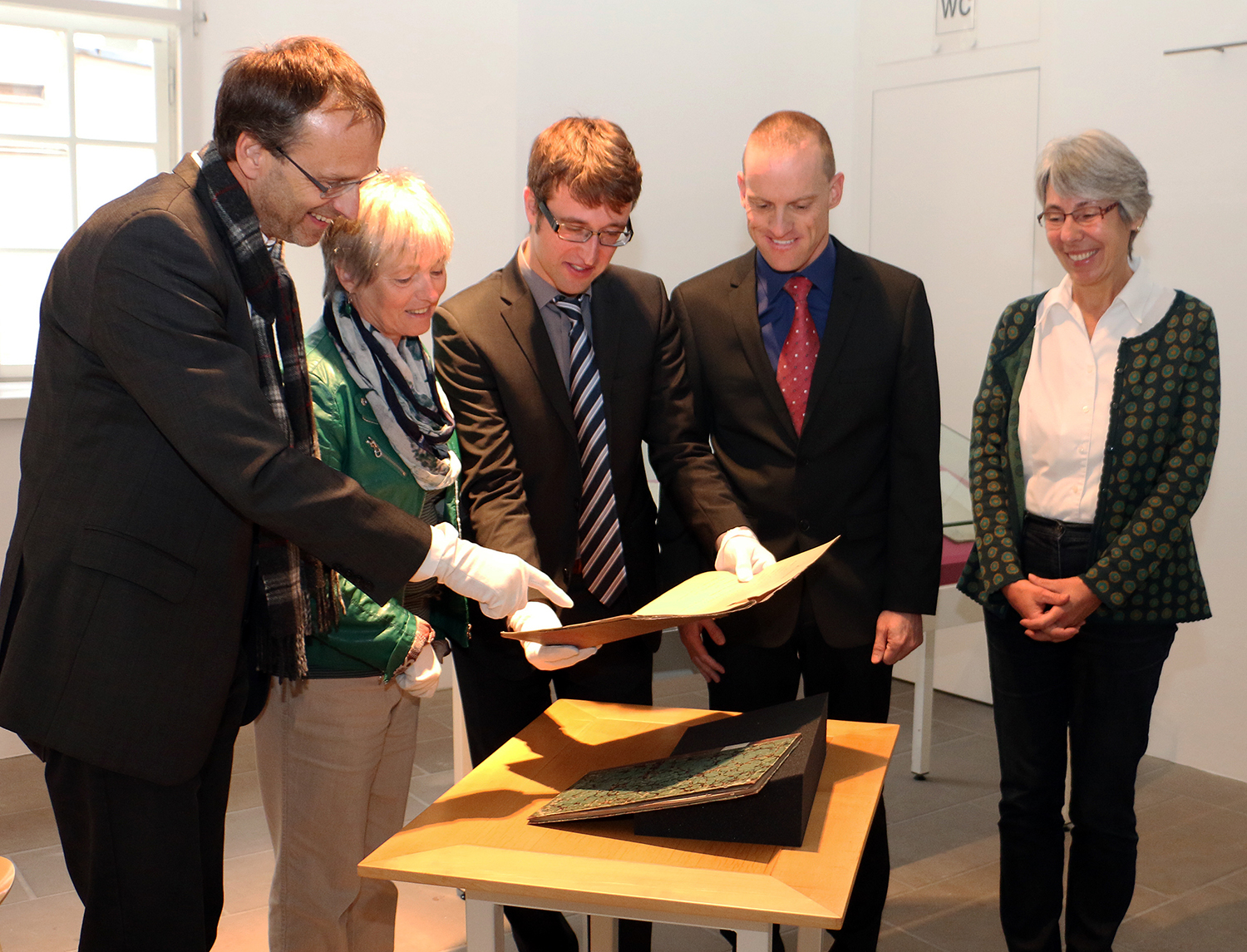 Fünf Personen stehen an einem Holztisch in der Hochschule Coburg und betrachten ein historisches Dokument. Zwei von ihnen tragen Handschuhe und halten das Dokument in der Hand, während die anderen zuschauen. Sie befinden sich in einem gut beleuchteten Raum mit einem Fenster, im Hintergrund ist ein "WC"-Schild zu sehen.