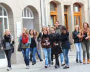 Eine Gruppe junger Leute, möglicherweise Studenten der Hochschule Coburg, schlendert eine Kopfsteinpflasterstraße entlang. Sie lächeln und halten Blumen in den Händen. Die Straße ist gesäumt von Gebäuden mit gewölbten Fenstern und Türen. Einige tragen Jacken und Schals.