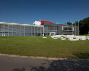 Vor dem klaren blauen Himmel der Hochschule Coburg erhebt sich ein modernes Gebäude mit Glasfassade und geometrischem Design. Es ist von einer grünen Wiese umgeben, im Vordergrund stehen weiße Steinbänke, im Hintergrund Bäume, die eine harmonische Verbindung aus Natur und Innovation schaffen.