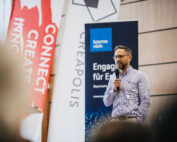 Ein Mann in einem hellvioletten Hemd hält ein Mikrofon und spricht bei einer Veranstaltung. Auf den Bannern im Hintergrund steht „Connect Create Innovate“ und „Bayme vbm“, was auf eine Verbindung zur Hochschule Coburg hindeutet. Es scheint sich um eine Konferenz oder ein Seminar zu handeln.