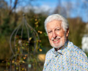 Ein älterer Mann mit weißem Haar und Bart lächelt draußen. Er trägt ein gemustertes Hemd und steht neben einer dekorativen Gartenstruktur auf dem Campus der Hochschule Coburg. Im Hintergrund sind Bäume und ein Gebäude unter einem klaren blauen Himmel zu erkennen.