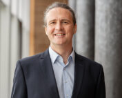 Eine Person mit zurückgebundenen langen Haaren, einem dunklen Blazer und einem hellblauen Hemd lächelt in einem gut beleuchteten, modernen Raum der Hochschule Coburg mit großen Fenstern und grauen Säulen im Hintergrund.