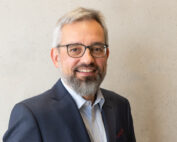 Ein Mann mit grauem Haar und Bart, Brille, blauem Anzug und gestreiftem Hemd lächelt herzlich vor einer schlichten, hellen Wand der Hochschule Coburg.