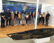 Eine Gruppe von Menschen mit Mundschutz steht in der Galerie der Hochschule Coburg. Hinter ihnen hängt ein großes Gemälde mit einer Meeresszene an der Wand. Im Vordergrund ist eine Kunstskulptur ausgestellt, die an ein Boot erinnert.