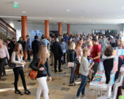 Eine große Gruppe von Menschen hat sich bei einer gut besuchten Veranstaltung in der Hochschule Coburg versammelt. Die Teilnehmer gehen umher, unterhalten sich und stöbern an verschiedenen Informationsständen und Displays. Einige halten Broschüren in der Hand. Der Raum ist gut beleuchtet, natürliches Licht fällt durch die Fenster.