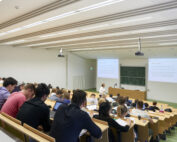 Der Hörsaal der Hochschule Coburg ist voller Studenten, die in Tischreihen sitzen und aufmerksam nach vorne blicken, wo ein Dozent einen Vortrag hält. Auf zwei Projektionsflächen und einer großen Tafel werden Notizen und Diagramme angezeigt, was das akademische Erlebnis noch bereichert.
